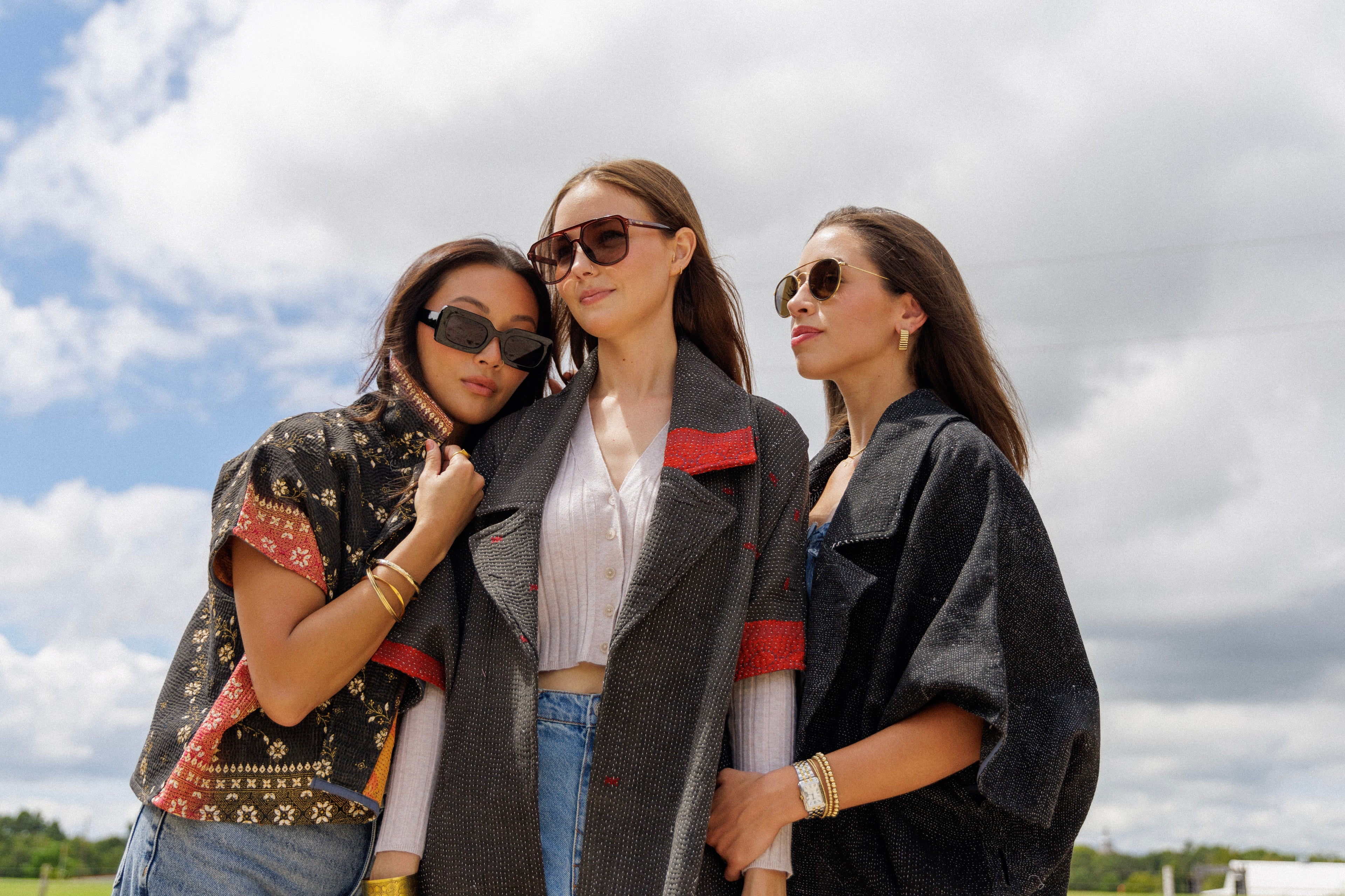 Three women standing outdoors under a cloudy sky, styled in coordinated, fashion-forward looks. They’re wearing statement outerwear pieces with a relaxed, oversized fit—featuring short wide sleeves, textured fabrics, and bold detailing like red accents and intricate patterns.